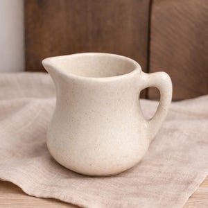 May include: A small, speckled cream-colored ceramic creamer with a rounded body and a curved handle. The creamer has a spout for easy pouring and sits on a beige linen napkin. The background features a wooden surface.