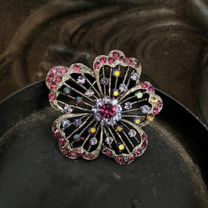 May include: A flower-shaped brooch with a dark metal frame. The brooch is adorned with pink and purple rhinestones, with a larger pink stone at the center. The petals are outlined with pink stones, and the center has a cluster of smaller, colorful stones.