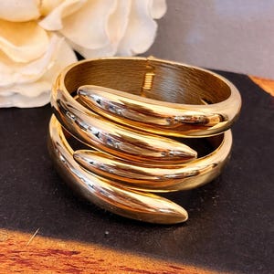 May include: A gold-colored bracelet with a unique design. The bracelet wraps around the wrist with a smooth, polished finish. The bracelet is a statement piece, perfect for adding a touch of elegance to any outfit.