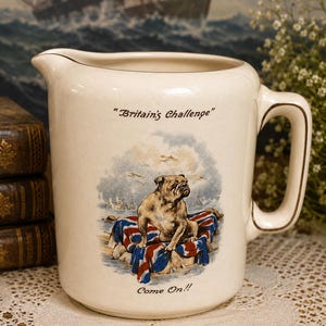 Puede incluir: Una jarra de cerámica de color crema con una ilustración central de un bulldog sentado sobre una bandera Union Jack. El texto "Britain's Challenge" está impreso sobre el perro y el texto "Come On!!" aparece debajo de la ilustración.