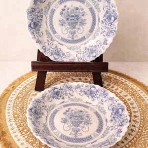 May include: Two white ceramic plates with a blue floral design. The plates have a scalloped edge and are displayed on a wooden easel and a woven placemat. The floral pattern includes roses and a central bouquet.
