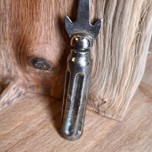 May include: A vintage, silver-toned bottle opener with a decorative top. The opener has a long, ridged handle and a pointed end with a curved, forked design. The background features a wooden surface.