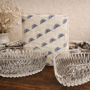 Puede incluir: Dos cuencos de vidrio transparente estampados y un paquete envuelto con un diseño floral azul. Los cuencos tienen un diseño acanalado y están sobre una superficie de madera. Un jarrón de flores blancas está en el fondo.