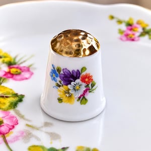 May include: A white porcelain thimble featuring a metallic gold-toned hammered top and a colourful floral bouquet design on the side, displaying purple, yellow, orange, blue, and white flowers with green leaves against a smooth white surface.