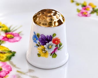 Peut inclure: Un dé à coudre en porcelaine blanche avec un sommet martelé de couleur or métallique et un motif de bouquet floral coloré sur le côté, montrant des fleurs violettes, jaunes, orange, bleues et blanches avec des feuilles vertes sur fond blanc.