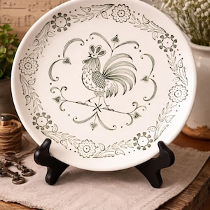 May include: White ceramic plate with a green rooster illustration in the center, surrounded by a floral and vine border. The plate is displayed on a black stand, showcasing a vintage design.