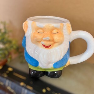 May include: A ceramic mug shaped like a garden gnome. The mug features a white beard, orange skin, and a blue jacket. The handle is white, and the gnome's feet are black. The mug is designed with a smiling face and closed eyes.