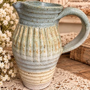 May include: A ceramic pitcher featuring a light blue and cream glaze with brown speckles. The upper section has vertical fluted ridges, while the lower portion displays horizontal ribbed textures. It includes a curved handle and a spout.