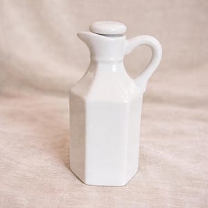 Puede incluir: Una aceitera de cerámica blanca con tapa, asa y boquilla. La aceitera tiene una forma geométrica octogonal. El asa es curva y la boquilla es pequeña e inclinada. La tapa es redonda y se coloca encima.