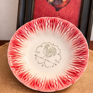 May include: A decorative ceramic plate with a red and white scalloped rim. The center features a rooster illustration in a silver color, surrounded by a floral design. A framed rooster painting is in the background.