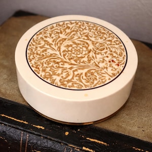 May include: A round, off-white decorative box with a detailed floral pattern in shades of brown on the lid. The lid is bordered by a thin black line. The box sits on a dark surface, suggesting a vintage or antique aesthetic.