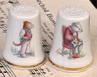 Peut inclure: Deux dés à coudre en porcelaine blanche avec des bordures dorées, chacun orné d'une illustration fantaisiste. L'un représente une souris en gilet, l'autre un blaireau en manteau. Les dés à coudre sont posés sur une partition de musique ancienne.
