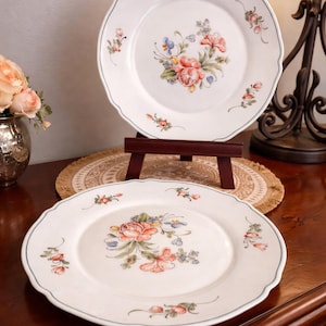 May include: Two white ceramic plates with scalloped edges and a floral design. The plates feature a central bouquet of pink, orange, and blue flowers, with additional floral accents around the rim. A blue line borders the edge.