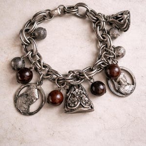 Puede incluir: Pulsera de abalorios plateada con un diseño de cadena gruesa. La pulsera está adornada con varios abalorios, incluyendo cuentas redondas, un abalorio en forma de campana y colgantes circulares con diseños en relieve. Los abalorios son una mezcla de plata y marrón oscuro.