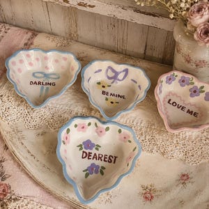 May include: Four heart-shaped ceramic dishes with hand-painted designs. One features pink polka dots and a blue bow with the text DARLING. Another has yellow flowers and a purple bow with BE MINE. One says LOVE ME with purple flowers. One says DEAREST with pink and purple flowers.