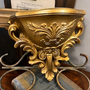 May include: Ornate gold-colored wall shelf with intricate scrollwork and a fleur-de-lis design. The shelf is supported by a decorative metal bracket. A vintage book and two metal rings are in the foreground.