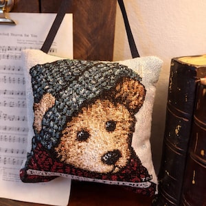 May include: A small decorative pillow featuring a teddy bear wearing a blue hat and a red scarf. The pillow has a neutral background and a dark brown strap for hanging. The image is a close-up of the pillow.