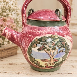 May include: A round ceramic teapot with a high arched handle and a matching lid. The body features a mottled magenta glaze with a central circular hand-painted scene of a tree in a landscape, framed by green leaves, set against a green base.