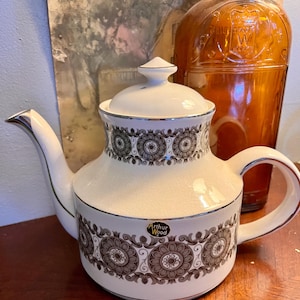 Puede incluir: Tetera de cerámica color crema con un estampado floral en marrón y gris. La tetera tiene un ribete plateado y una tapa con un pequeño pomo. El asa y el pico son curvos. La etiqueta dice "Arthur Wood".