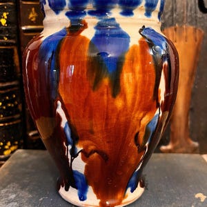 Puede incluir: Un jarrón de cerámica con un esmalte único. El jarrón presenta una base blanca, con una mezcla de tonos marrones, azules y naranjas. El borde es azul oscuro. Los colores gotean por los lados, creando un efecto texturizado. Es un objeto decorativo.