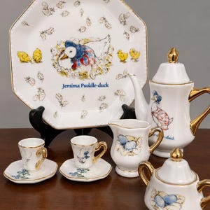 Può includere: Un servizio da tè in porcellana bianca con finiture dorate, con illustrazioni di un'anatra e anatroccoli. Il set include un piatto, una teiera, una lattiera, una zuccheriera e due tazze con piattini. Il piatto presenta la scritta "Jemima Puddle-duck".