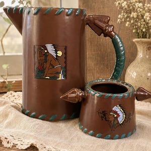 May include: A brown ceramic pitcher and a matching smaller vessel with a handle. Both items feature a teal stitched-look border and graphic illustrations of a person wearing a feathered headdress, with the pitcher showing a kneeling figure.