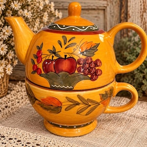 May include: A ceramic tea for one set featuring a yellow teapot stacked on a matching teacup. The teapot is decorated with hand-painted red apples, grapes, and green leaves. The teacup features a matching painted vine pattern around the exterior.