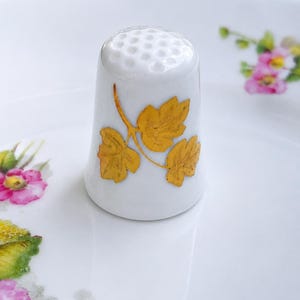 Peut inclure: Un d&eacute; &agrave; coudre en porcelaine blanche avec un sommet textur&eacute; et un motif central compos&eacute; de trois feuilles jaune dor&eacute; sur une tige fine. Le d&eacute; est pos&eacute; sur une surface blanche orn&eacute;e de petits motifs floraux roses et jaunes.