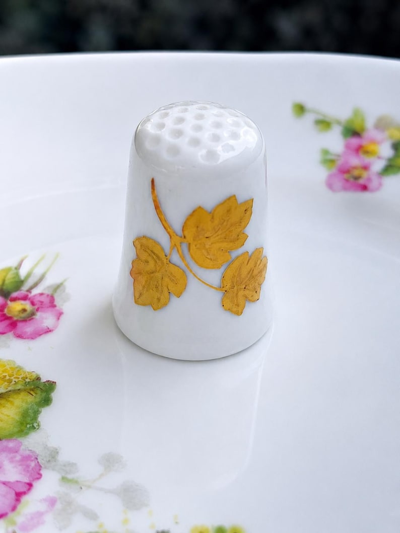Peut inclure: Un d&eacute; &agrave; coudre en porcelaine blanche avec un sommet textur&eacute; et un motif central compos&eacute; de trois feuilles jaune dor&eacute; sur une tige fine. Le d&eacute; est pos&eacute; sur une surface blanche orn&eacute;e de petits motifs floraux roses et jaunes.