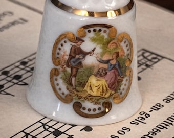 Peut inclure: Dé à coudre en porcelaine blanche avec un bord doré et des accents dorés décoratifs. Le dé à coudre présente une scène peinte d'un homme jouant de la guitare pour deux femmes. Le dé à coudre repose sur une partition.