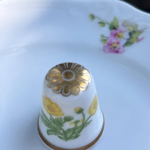 May include: A white porcelain thimble featuring a gold-painted flower design on the top and yellow floral illustrations on the side. The thimble has a thin gold band around the base and sits on a white porcelain saucer with a small pink floral motif.