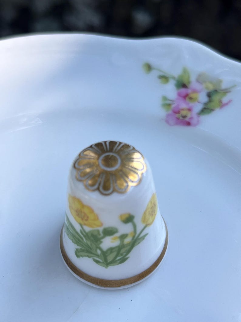 May include: A white porcelain thimble featuring a gold-painted flower design on the top and yellow floral illustrations on the side. The thimble has a thin gold band around the base and sits on a white porcelain saucer with a small pink floral motif.
