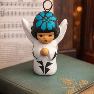 Puede incluir: Figura de ángel de cerámica con alas blancas y un diseño floral negro y azul en la cabeza. El ángel junta las manos en señal de oración. La figura está sobre un libro antiguo.