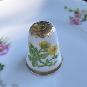 May include: A white porcelain thimble featuring a hand-painted yellow buttercup flower design with green leaves. The top of the thimble is decorated with a gold-colored flower motif, and the base is finished with a thin gold-colored band.