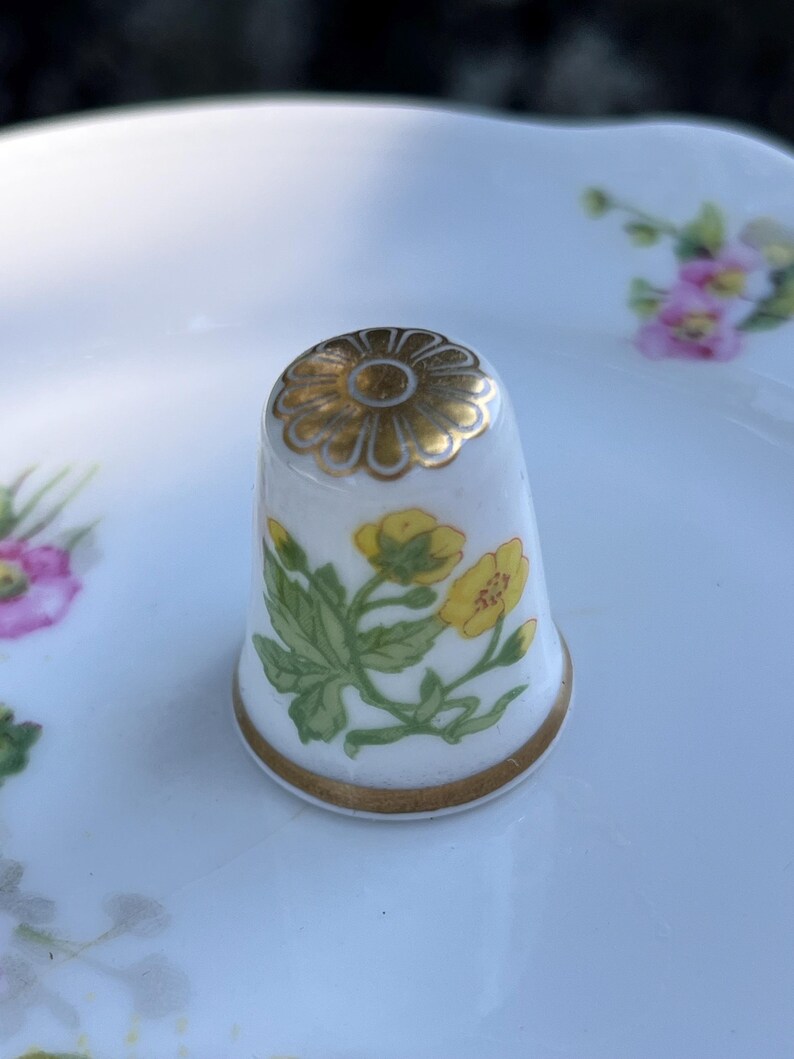 May include: A white porcelain thimble featuring a hand-painted yellow buttercup flower design with green leaves. The top of the thimble is decorated with a gold-colored flower motif, and the base is finished with a thin gold-colored band.