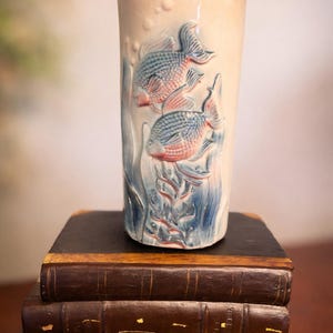Puede incluir: Un jarrón de cerámica color crema con forma cilíndrica, con peces azules y rojos en relieve nadando entre plantas acuáticas. El jarrón se exhibe sobre una pila de tres libros antiguos encuadernados en cuero.