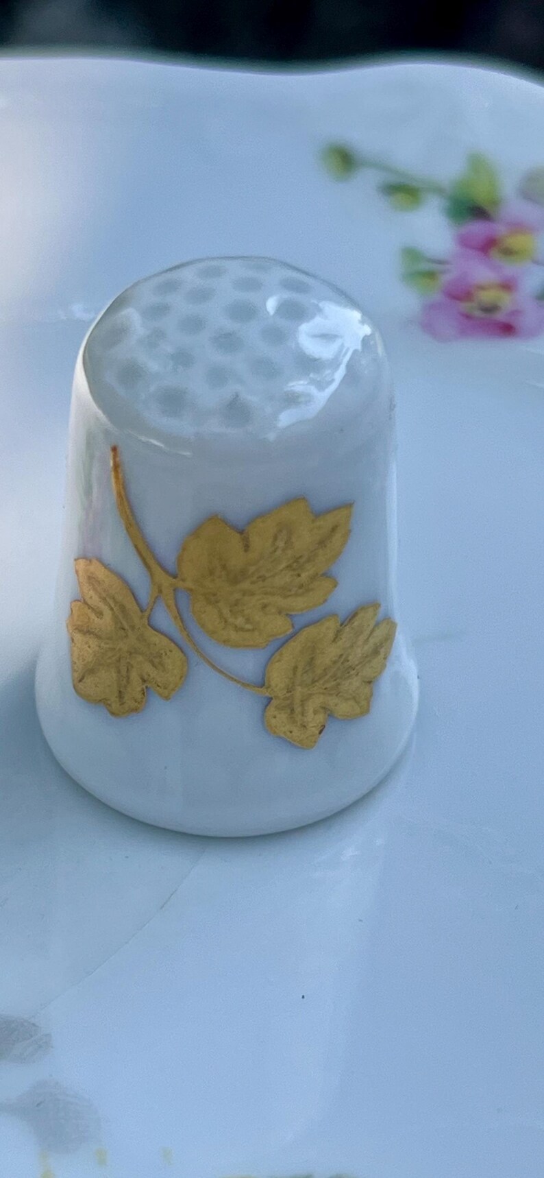 Peut inclure: Un d&eacute; &agrave; coudre en porcelaine blanche avec un sommet textur&eacute; alv&eacute;ol&eacute; et un motif de feuilles dor&eacute;es sur le c&ocirc;t&eacute;. Le d&eacute; pr&eacute;sente une base &eacute;vas&eacute;e et une finition lisse et brillante, orn&eacute; d'une illustration dor&eacute;e de trois feuilles reli&eacute;es.