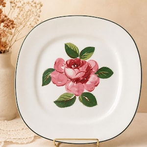 May include: A square white ceramic plate with rounded corners and a thin dark green rim. The center of the plate features a painted illustration of a large pink rose with several green leaves, displayed on a gold metal stand.