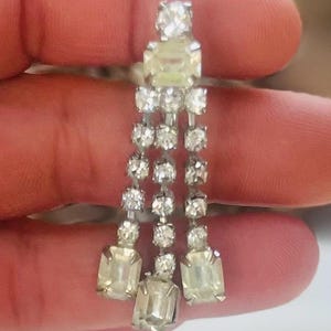 以下が含まれることがあります： クリアでキラキラ光るラインストーンが連なった、シルバーカラーのぶら下がりピアス。ピアスは、上部に大きな長方形の石、その下に小さな丸い石が3列、下部に長方形の石が3つ付いています。