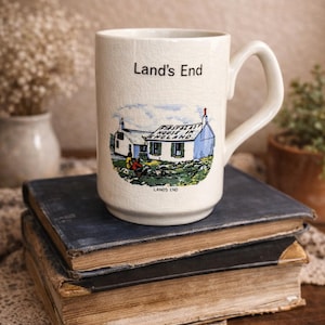 May include: Cream-colored ceramic mug with a handle, featuring a colorful illustration of a building with the text "Land's End" and "First & Last House in England." The mug is placed on a stack of old books.