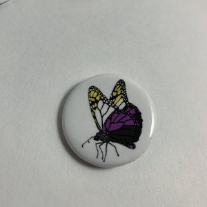 May include: A white button with a purple, yellow, and black butterfly design. The butterfly is in flight with its wings spread.