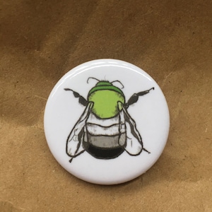 May include: A white button with a black and green bee illustration. The bee has a green head and thorax, black abdomen, and white wings.
