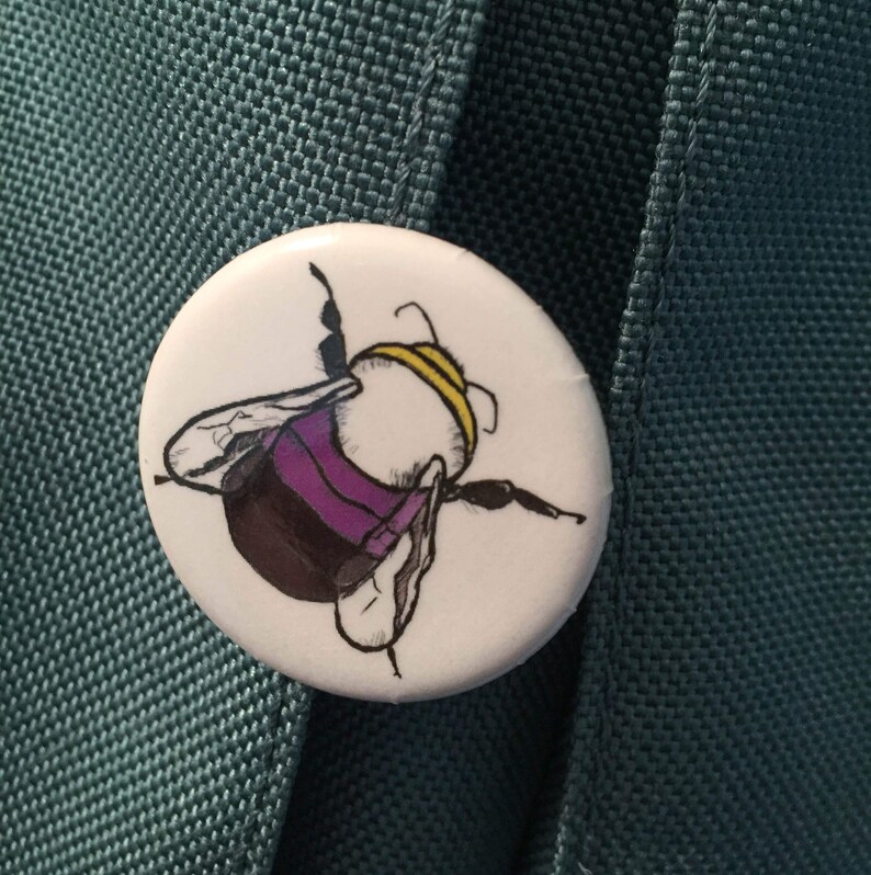 May include: A white button with a black and purple bumblebee illustration. The bumblebee has yellow stripes on its abdomen and is facing away from the viewer.