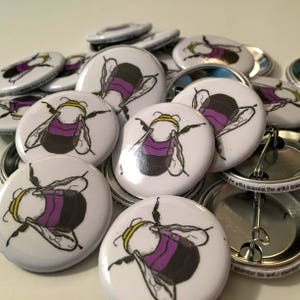 May include: A close-up of a pile of round, white button pins with a black and purple bee illustration. The bee has yellow accents and is drawn in a cartoon style.