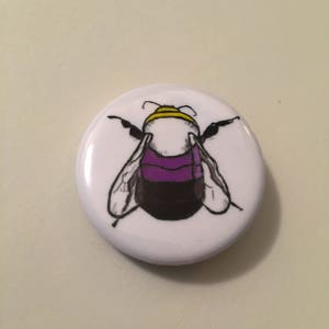 May include: A white button with a black and yellow bumblebee with purple stripes on its abdomen.