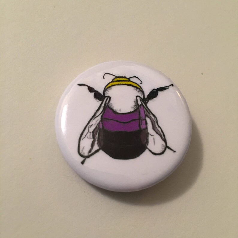 May include: A white button with a black and yellow bumblebee with purple stripes on its abdomen.