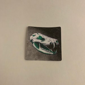 Peut inclure: Un autocollant représentant un crâne vert et blanc avec des dents sur un fond gris foncé avec un motif texturé.