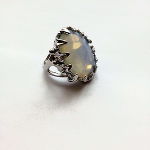 May include: A silver ring featuring a large, oval-shaped, opalescent gemstone. The gemstone is surrounded by a decorative, textured silver setting. The ring is set against a white background, highlighting its design and color.