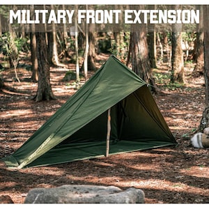 May include: A green military front extension tent set up in a woodland environment. The tent is propped up with a wooden pole and features the text "MILITARY FRONT EXTENSION". The ground is covered in pine needles.