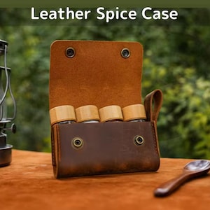 Peut inclure: Un étui à épices en cuir marron avec le texte "Leather Spice Case" en haut. L'étui est ouvert, révélant quatre pots à épices en verre avec des couvercles en bois. Une cuillère en bois repose sur la surface.